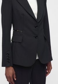 Blazer noir sur mesure avec deux boutons, poches à rabat, fermetures éclair décoratives et boutons aux manches, porté sur un haut blanc avec un pantalon noir.