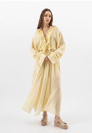Femme portant une robe fluide jaune pâle avec un décolleté en V à volants et de larges poignets, associée à des sandales compensées beige, debout devant un fond blanc.