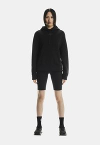 Schwarzer Kapuzenpullover mit Kängurutasche, kombiniert mit schwarzen Shorts bis zur Mitte des Oberschenkels und schwarzen Sneakers. Minimalistisches Design, weiche Textur.