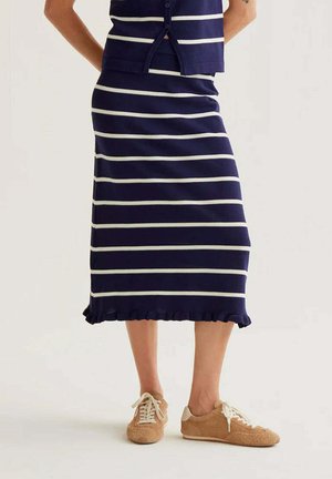 Persona que lleva una falda azul marino con rayas horizontales blancas y dobladillo con volantes, combinada con zapatos casuales beige con cordones sobre un fondo blanco.