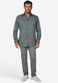 Blouse met lange mouwen in donkerblauw met een bloemenpatroon, gecombineerd met grijze slim-fit broek en witte sneakers. Effen achtergrond.