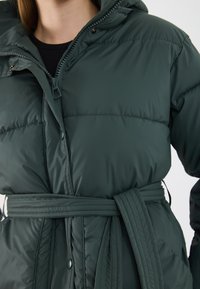 Donkergroene pufferjack met een modal textuur, een capuchon, een riem in de taille en zichtbare stikseldetails met drukknoop- en ritssluiting.
