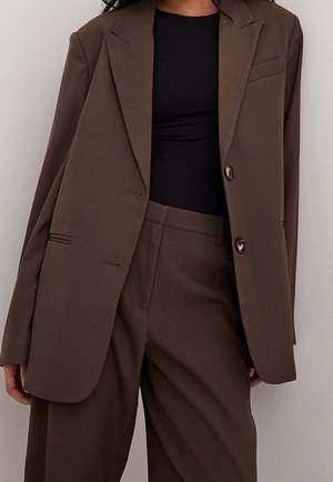 Nainen pukeutuneena ruskeaan ylisuurikokoiseen bleiseriin ja vastaaviin leveälahkeisiin housuihin, mustan istuvan topin päällä, seisomassa vaaleaa seinää vasten.