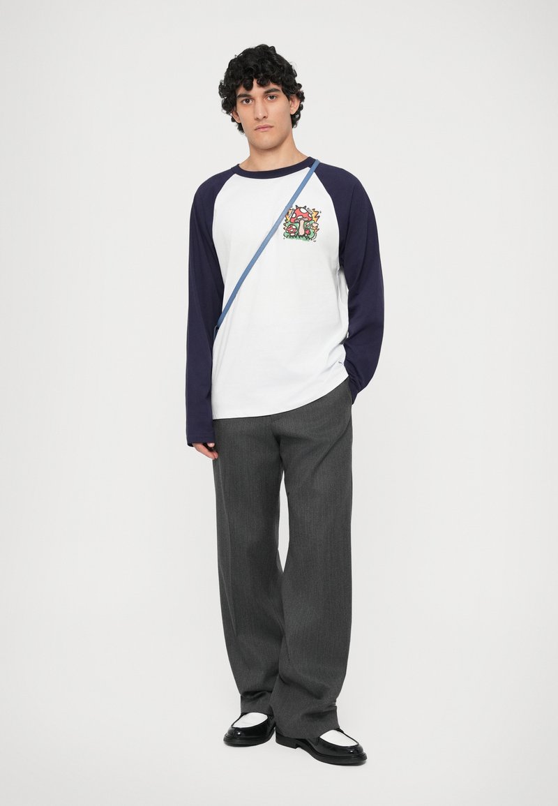 T-shirt à manches longues blanc et marine avec un graphisme vibrant sur la poitrine, associé à un pantalon large gris et des chaussures noires avec des accents blancs.