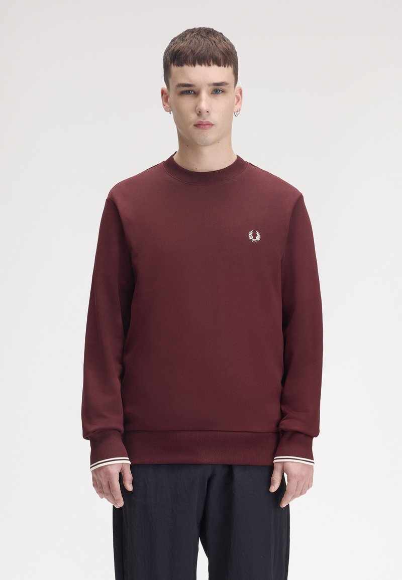Jeune homme portant un sweat bordeaux avec un petit logo blanc sur la poitrine et un pantalon noir, se tenant devant un fond uni.