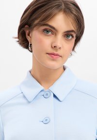 Lichtblauw overhemd met kraag en twee knoopsluitingen, met een gladde textuur en een getailleerde pasvorm. Korte mouwen zichtbaar aan de randen.