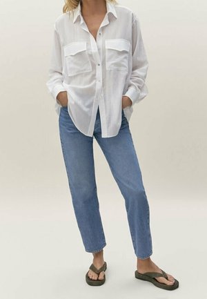Kvinde iført en stor hvid skjorte med knapper, blå jeans med lige ben og olivengrønne flipflops, stående med hænderne i lommerne.