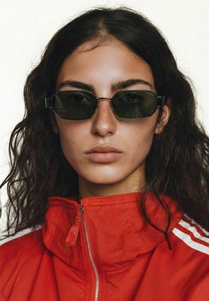 Jeune personne aux longs cheveux bouclés et foncés, portant des lunettes de soleil rectangulaires noires et une veste zippée rouge avec des rayures blanches.