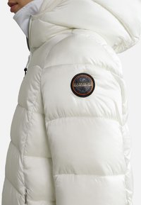 Biała kurtka puchowa z zapięciem na zamek błyskawiczny z przodu, kapturem i przeszywanymi panelami. Posiada okrągłą naszywkę z logo na lewym ramieniu. Gładka tekstura.