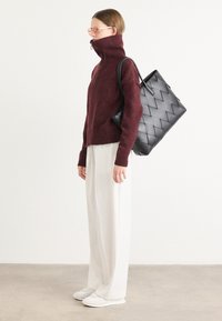Bordeaux hoge hals trui met geribbelde manchetten, gecombineerd met crèmekleurige wijde broek en witte sneakers. Zwarte geweven tote bag hangt over de schouder.