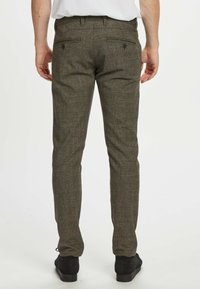 Tumši zaļi tekstūrēti bikses ar šauru piegriezumu un divām aizmugurējām kabatām, kombinētas ar melnām kurpēm, skatīts no aizmugures.