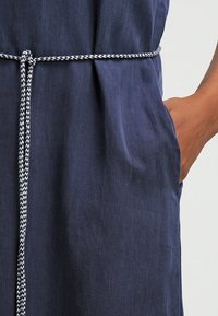 Personne portant une robe bleu foncé avec une ceinture tressée blanche et noire nouée à la taille, main placée dans la poche droite.