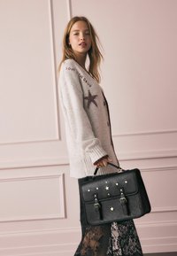 Czarna skórzana torebka z złotymi gwiazdkami, o solidnym kształcie, z podwójnymi paskami zapinanymi na sprzączki, trzymana w dłoni; zestawiona z przytulnym jasnym swetrem.