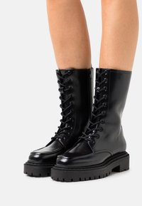 Bottes basses en cuir noir avec une finition brillante, dotées d'un laçage avant, d'une semelle robuste et d'une semelle extérieure texturée pour une meilleure adhérence. Tige lisse et bout arrondi.