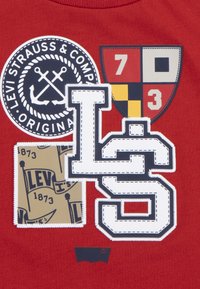 Vörös szövet több folttal: egy kör alakú logó horgonyokkal, két címer számmal és egy bézs folt zászlókkal és szöveggel.