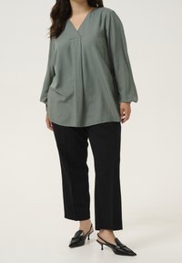 Blouse vert clair avec un col en V, motif à pois texturé, manches longues ; associée à un pantalon large noir et des mules à talons noires.