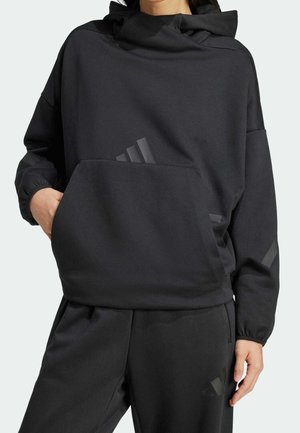 Persoon draagt een losse zwarte hoodie en bijpassende zwarte broek met elastische boorden en subtiele donkere strepen op de mouw en broekspijp.