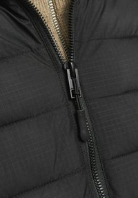 Chaqueta negra acolchada con una cremallera suave y un tirador texturizado, sobre un fondo de tela de punto beige. Patrón de cuadrícula sutil en la tela.