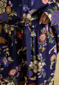 Cvjetne hlače s tamnoplavom pozadinom, koje imaju pojas s vezicom, složene uzorke u crvenoj, zelenoj i ljubičastoj boji, te glatku teksturu tkanine.