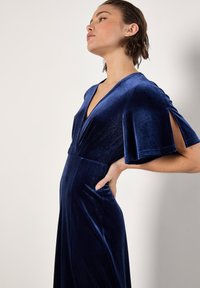 Robe en velours bleu marine avec un décolleté en V plongeant et des manches courtes, présentant des côtés ouverts. Le tissu a une texture douce et un design fluide.