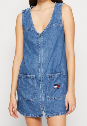 Kvinde iført ærmeløs blå denimkjole med lynlås foran og to forlommer, den ene med Tommy Jeans logo-patch.