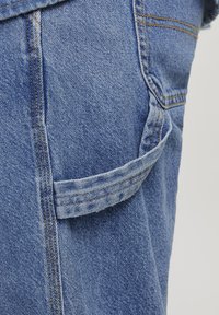 Tejido de denim en azul claro con costuras visibles. Presenta un bolsillo lateral con una solapa cosida, enfatizando un diseño casual y funcionalidad.