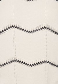 Pull en crème avec un motif en zigzag noir. Tissu tricoté texturé, avec des coutures décoratives et une légère brillance. Manches courtes.