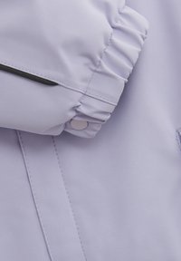 Ljuslila jacka med strukturerad tyg, samlad ärm med tryckknappsförslutning och kontrasterande svarta detaljer.