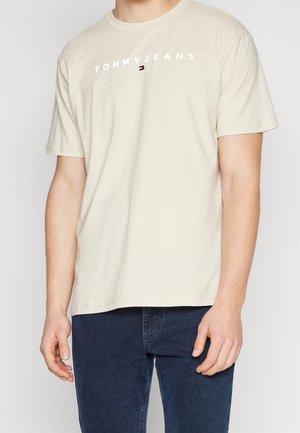 Mand iført beige Tommy Jeans T-shirt med korte ærmer og mørkeblå jeans, stående foran en ensfarvet baggrund.