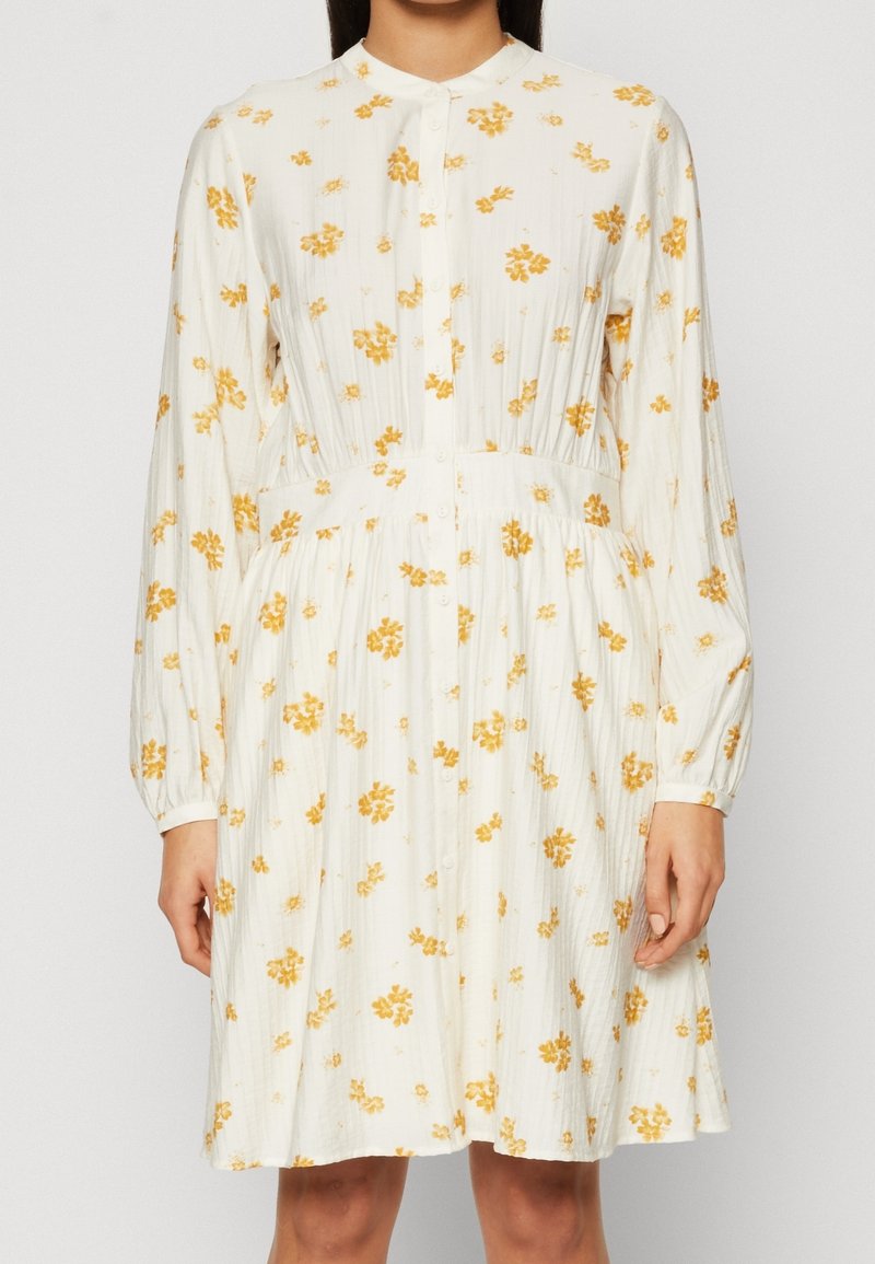 Femme portant une robe blanche à manches longues avec une patte de boutonnage sur le devant et un motif floral jaune, debout devant un fond uni.
