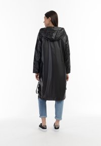 Zwarte regenjas met een capuchon, lange mouwen en verstelbare zijklappen. Gladde, glanzende textuur. Gecombineerd met opgerolde lichtblauwe jeans en donkere sneakers.