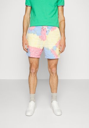 Mężczyzna ubrany w pastelowe spodenki tie-dye z białym sznurkiem, zieloną koszulkę polo, szare skarpetki i białe trampki, stojący na jednolitym tle.