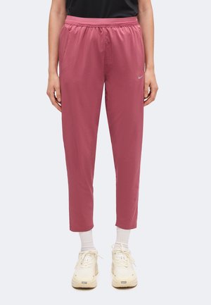 Persoon die roze Nike-joggingbroek, een zwart shirt, witte sokken en beige Nike-sneakers draagt, tegen een effen achtergrond.