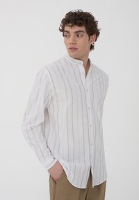 Camicia bianca a maniche lunghe con strisce verticali marrone chiaro, colletto alla cinese e chiusura a bottoni. Realizzata in tessuto leggero.