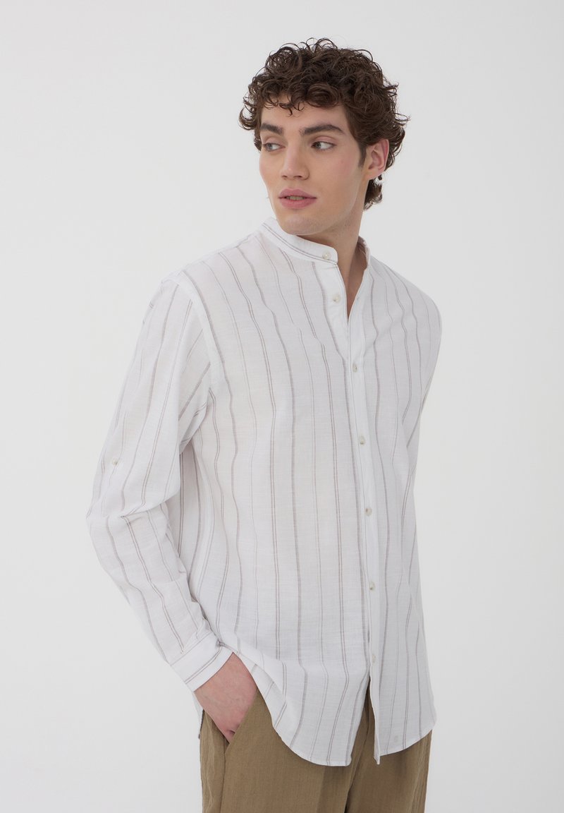 Camicia bianca a maniche lunghe con strisce verticali marrone chiaro, colletto alla cinese e chiusura a bottoni. Realizzata in tessuto leggero.
