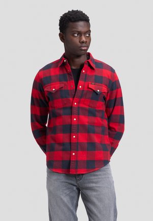 Giovane uomo con camicia a quadri rossa e nera e jeans grigi, che guarda di lato con le mani dietro la schiena su uno sfondo grigio uniforme.