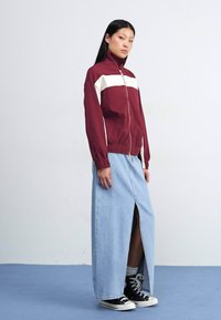 Kvinde står mod en lys baggrund, iført en bordeaux sportjakke, et langt denimskørt med en sideslits og sorte sneakers.