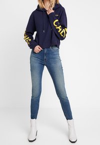 Kvinna i marinblå cropped hoodie med gul text på ärmarna, ljusblå skinny jeans och vita ankelboots, står med benen isär.