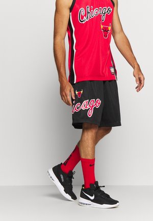 Mężczyzna ubrany w czerwoną koszulkę koszykarską Chicago Bulls, czarne spodenki z logo Chicago, czerwone skarpetki Nike i czarne buty sportowe Nike, stojący na szarym tle.