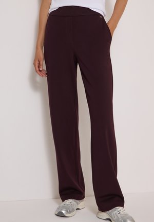 Personne portant un pantalon large violet foncé et des baskets argentées, debout contre un mur blanc, main dans la poche.