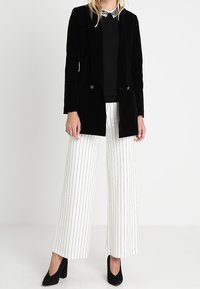 Blazer en velours noir à deux boutons, associé à un haut noir et un pantalon large rayé blanc, souligné par des escarpins noirs.