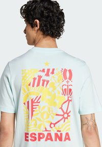 Camiseta de color azul claro con un gran gráfico colorido en la parte trasera, con patrones rojos y amarillos y la palabra "ESPAÑA" en una fuente roja en negrita.