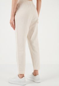 Light beige tapered pants with an elastic waistband and frayed hem, paired with white sneakers. Fabric appears soft and textured.