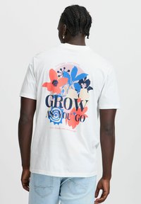 Hvit bomull t-skjorte med et fargerikt blomsterdesign og tekst på baksiden. Designet inkluderer rosa, blå og svarte elementer med en rund layout.