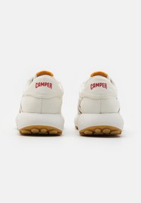 Λευκά αθλητικά παπούτσια με πορτοκαλί λεπτομέρεια στη γλώσσα, περιλαμβάνουν την κόκκινη επωνυμία "CAMPER", καουτσούκ σόλα και υφασμάτινο πάνω μέρος με υφή.