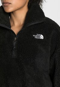 Maglione in pile nero con colletto alto e chiusura a zip. Presenta il logo "The North Face" ricamato in bianco sul petto. Texture morbida.