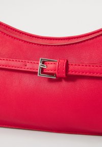 Rode leren handtas met een gladde textuur, gestructureerde vorm en een zilveren gesp die de brede schouderband accentueert.