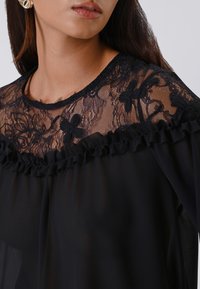Donna che indossa una blusa nera con un top in pizzo trasparente con ricami floreali e una cornice arricciata lungo il collo.