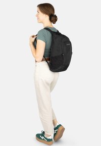 Schwarzer Stoff-Rucksack mit gepolsterten Trägern und einer Fronttasche, getragen über einem grauen Oberteil und cremefarbenen Hosen. Verfügt über eine glatte Oberfläche und ein stromlinienförmiges Design.