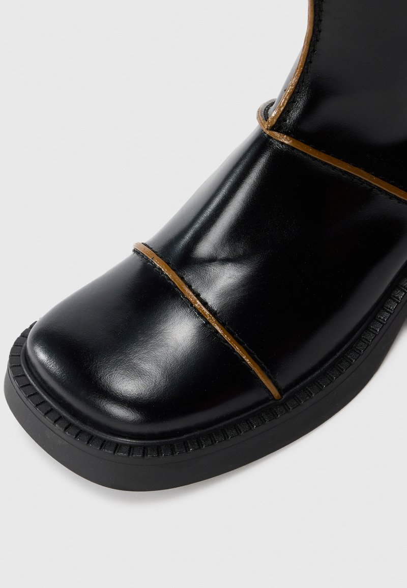 Svart läderstöveltå med tjock gummisula och bruna detaljer på den släta ytan, som visar en modern och robust design.